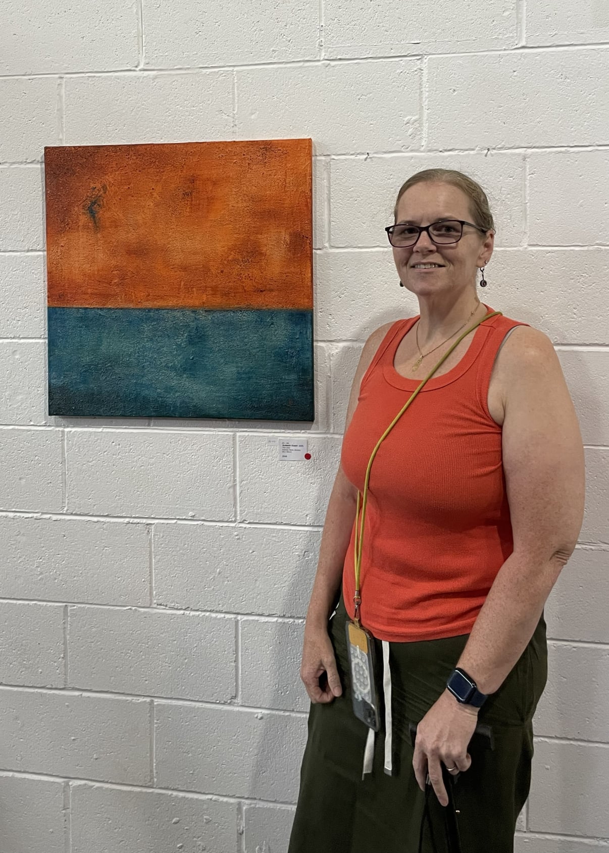 Artist Fiona Law wearing an orange top standing beside a square abstract painting with layered orange and blue textures